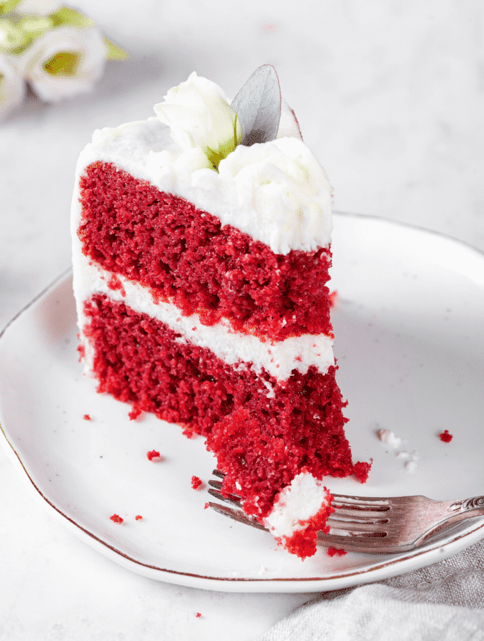 A piece of red velvet cake on a white plate. A fork is in front of the cake with a piece of cake on it.