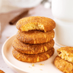 A stack of four snickerdoodle cookies on a white plate. The top cookie has a bite out of the front of it and there is a glass of milk directly to the right of the stack.