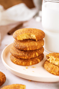 A stack of four snickerdoodle cookies on a white plate. The top cookie has a bite out of the front of it and there is a glass of milk directly to the right of the stack.