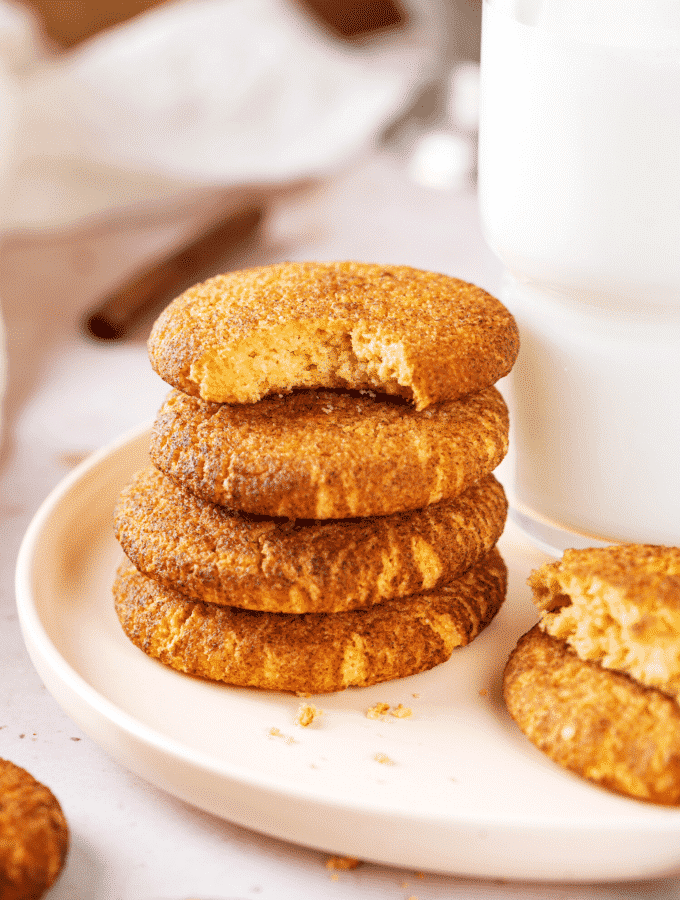 A stack of four snickerdoodle cookies on a white plate. The top cookie has a bite out of the front of it and there is a glass of milk directly to the right of the stack.