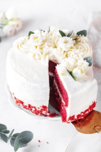 A red velvet cake on a white serving tray with a slice of cake being taken out of the front by a gold server.