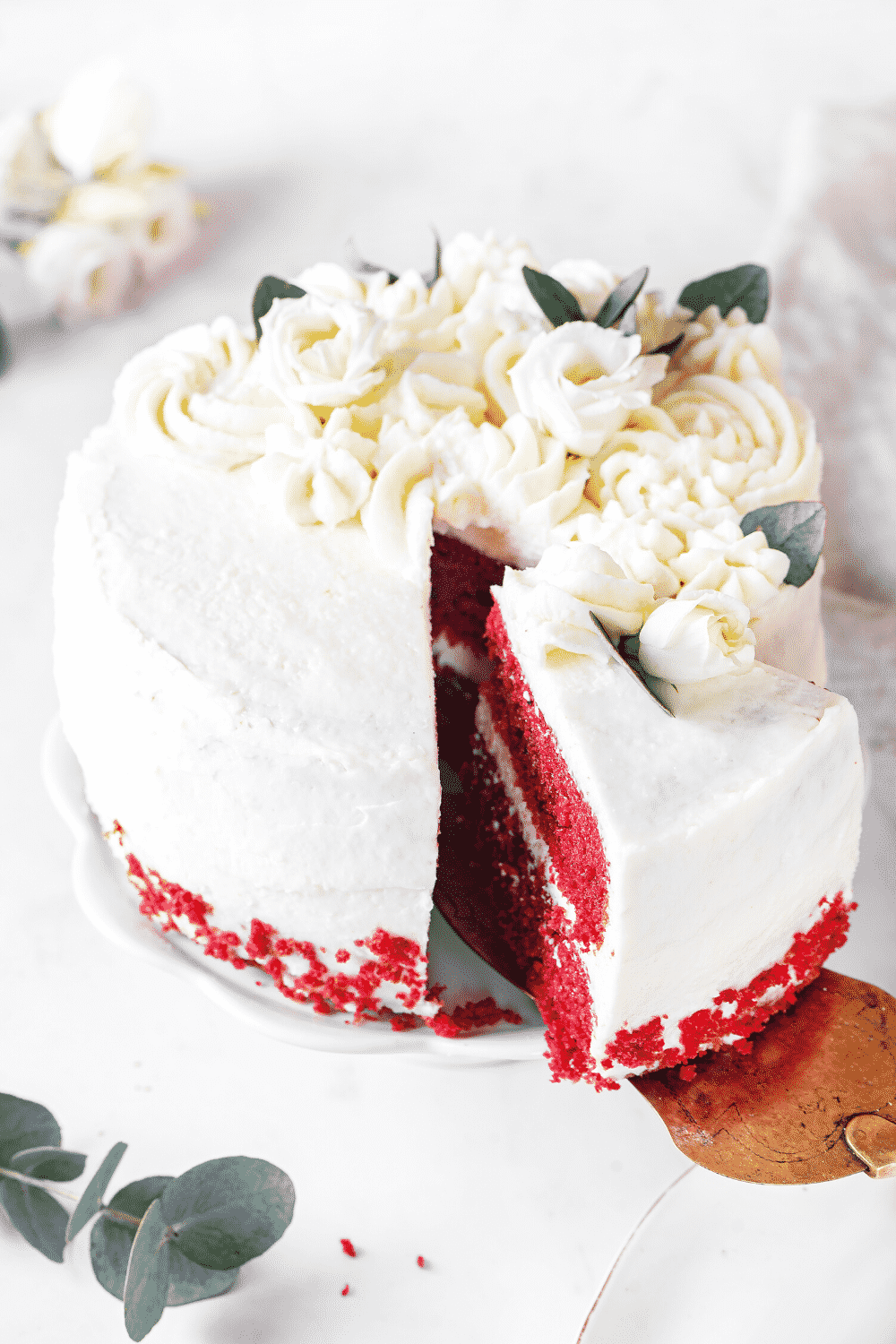 A red velvet cake on a white serving tray with a slice of cake being taken out of the front by a gold server.