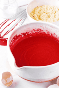 A white mixing bowl with red velvet cake batter in it. There is a white bowl behind it with the dry ingredients for the batter.