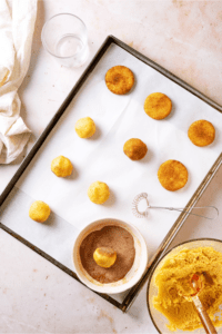 A baking sheet lined with parchment paper with with rows of snickerdoodle cookies on it. A few snickerdoodle cookies are rolled in cinnamon sugar coating and flattened and a few are still a dough ball. There is a bowl of cinnamon sugar on the parchment paper with one cookie in it.