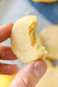 Two fingers holding a lemon cookie with a bite taken out of it.