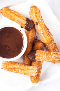 Seven churros overlapping one another on a piece of white parchment paper on a white plate. There is a cup of chocolate sauce to the left of the churros and two churros have chocolate sauce on the tip of them.