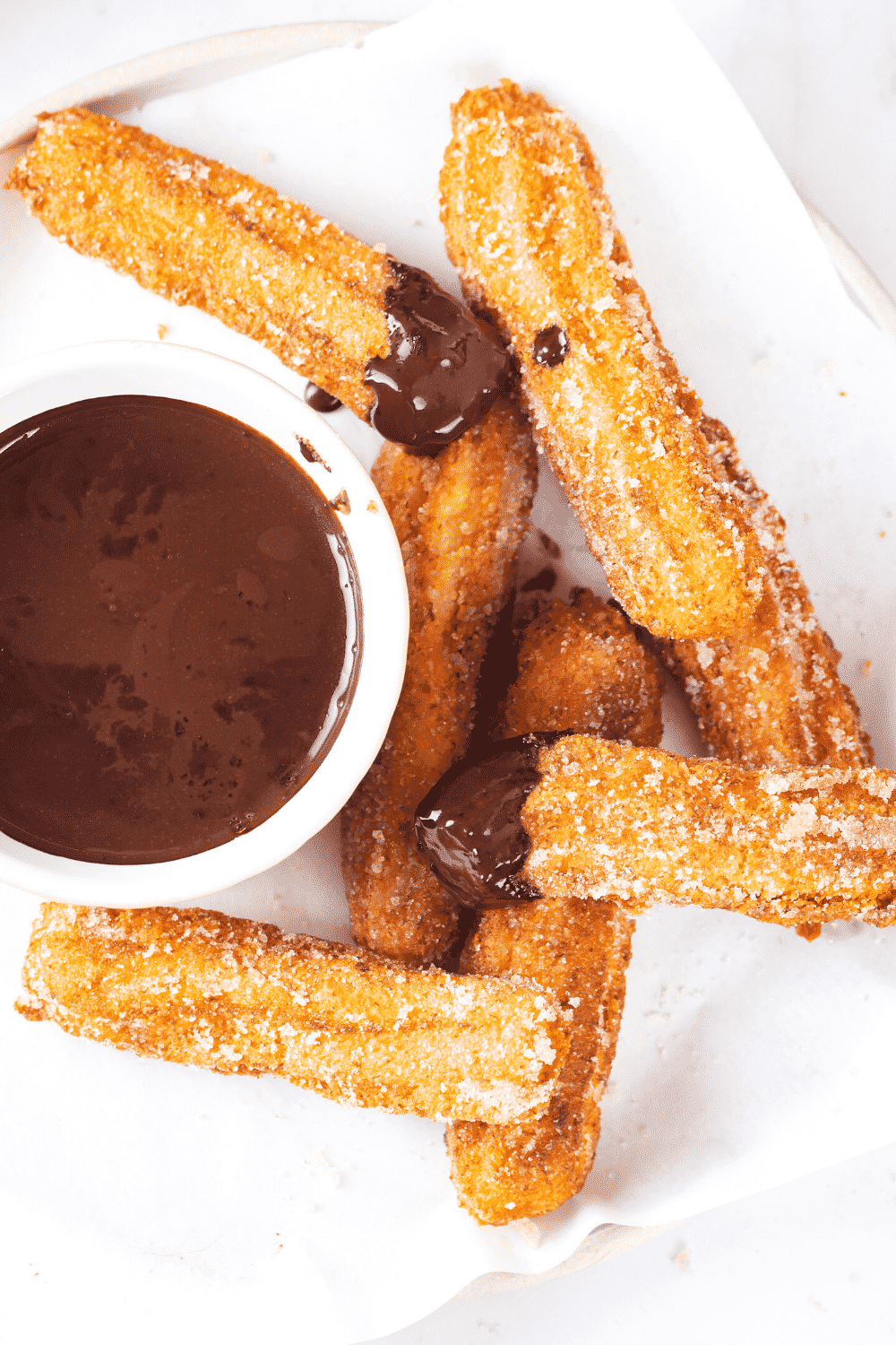Seven churros overlapping one another on a piece of white parchment paper on a white plate. There is a cup of chocolate sauce to the left of the churros and two churros have chocolate sauce on the tip of them.