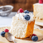 A piece of angel food cake on a plate. The piece of angel food cake has whipped cream and two blueberries and one raspberry on top.