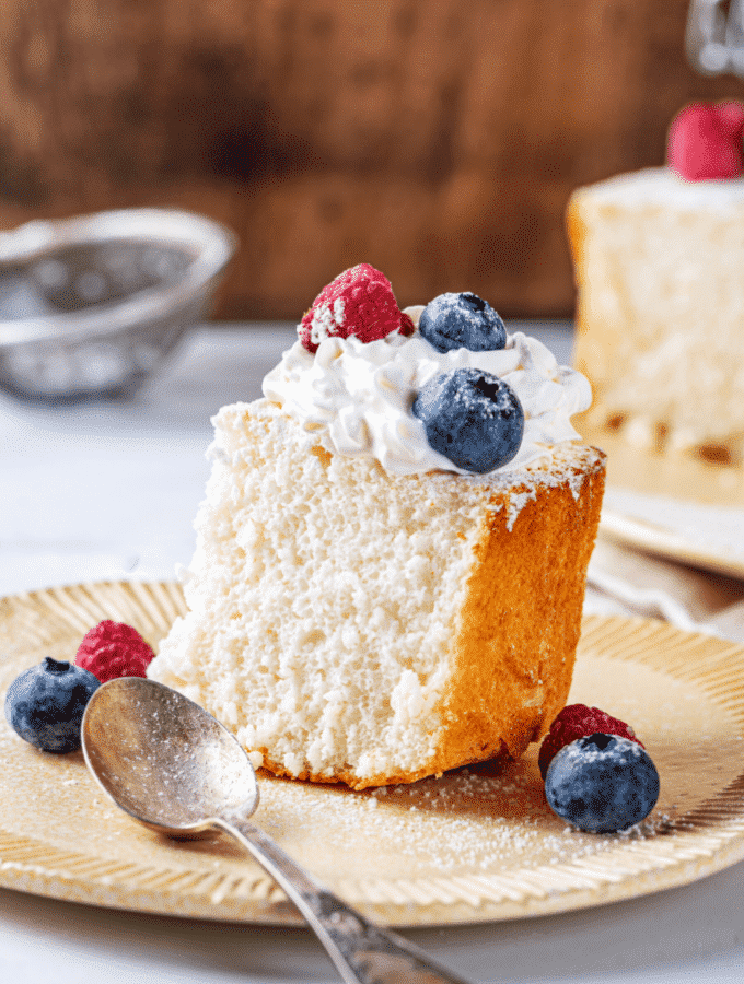 A piece of angel food cake on a plate. The piece of angel food cake has whipped cream and two blueberries and one raspberry on top.