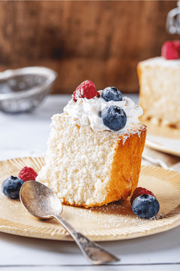 A piece of angel food cake on a plate. The piece of angel food cake has whipped cream and two blueberries and one raspberry on top.