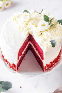 A red velvet cake on a white serving tray. A piece of cake is missing from the front of the cake.