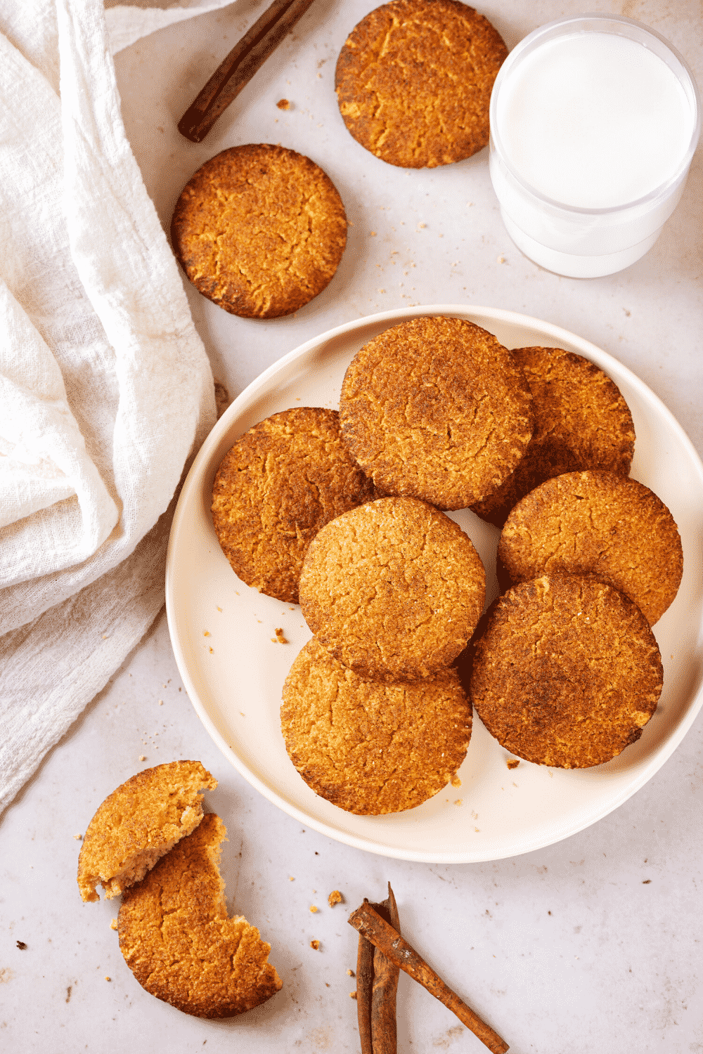 A white plate with several snickerdoodle cookies on it. There are a few snickerdoodle cookies on the white counter around the plate. Glass of milk is behind the plate of cookies.