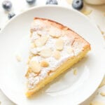 A piece of almond cake on a white plate. The plate is sat next to a cup of coffee and there are blueberries next to the cup and plate.