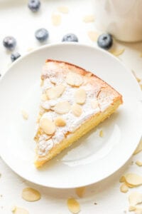 A piece of almond cake on a white plate. The plate is sat next to a cup of coffee and there are blueberries next to the cup and plate.