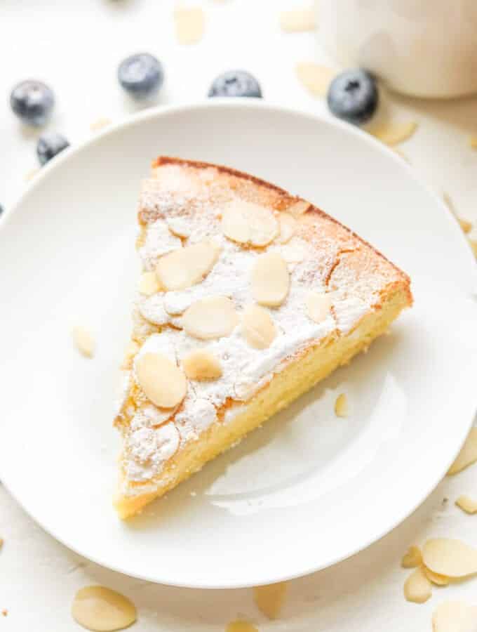 A piece of almond cake on a white plate. The plate is sat next to a cup of coffee and there are blueberries next to the cup and plate.