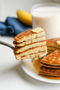 A fork holding a stack of cut up pancakes. The pancakes are topped with butter and there is a glass of milk, and a blue napkin behind them.