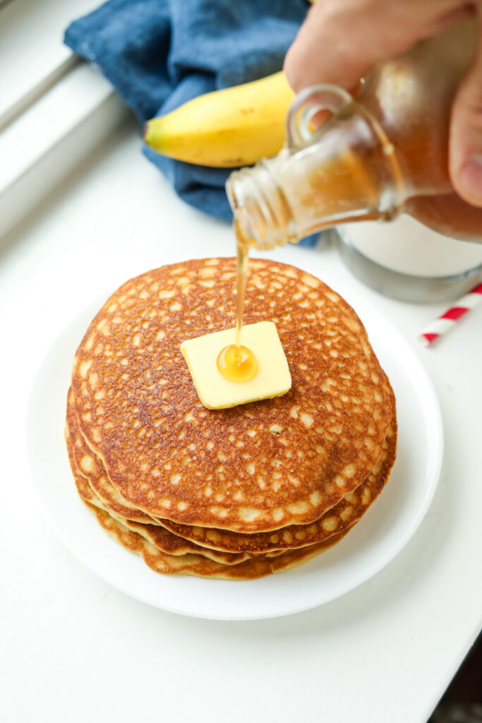 A hand holding a bottle of maple syrup, and pouring it on a stack of pancakes.