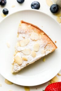 A slice of almond cake on a white plate. There are blueberries and a strawberry next to the plate.