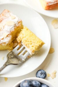 A piece of almond cake on a white plate with a fork holding a piece of the cake.