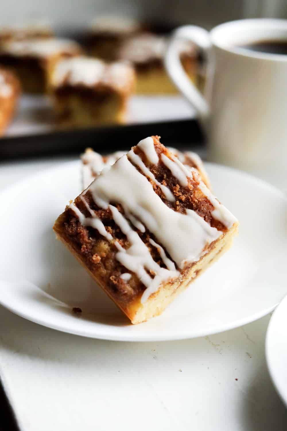 A white plate with a piece of coffee cake that is stacked on top of another piece of cake and a cup of coffee is set behind the plate. There are more pieces of cake next to the cup as well.