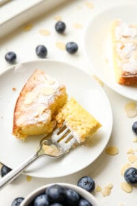 A slice of almond cake on a white plate, and a fork is holding a piece of the cake. There's a cup of blueberries next to the plate as well as some loose ones.