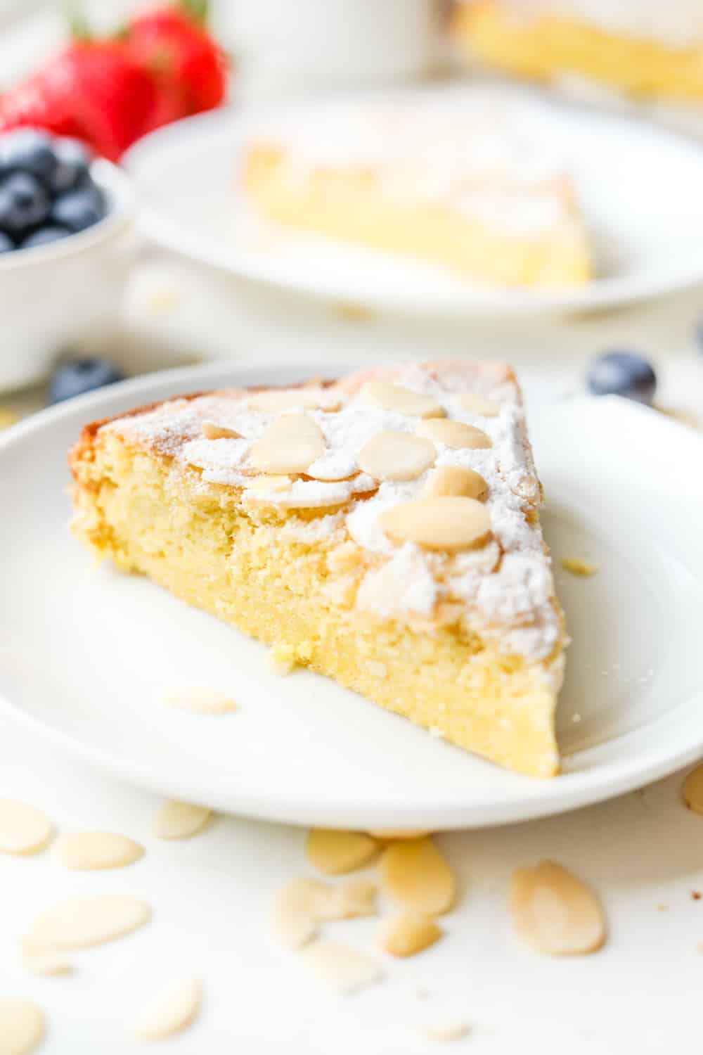 A slice of almond cake on a white plate. There's another slice of cake, a cup of blueberries, and some strawberries behind it as well.