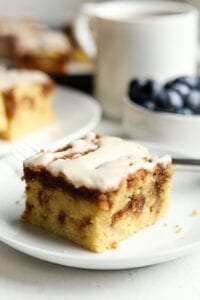 A piece of coffee cake on a white plate. There are more pieces in the background along with a cup of coffee and a bowl of blueberries.