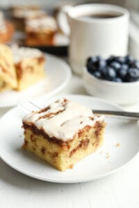 A piece of coffee cake on a white plate with a fork set by it. There's a coffee cup, blueberries, and more pieces of cake set behind the plate.