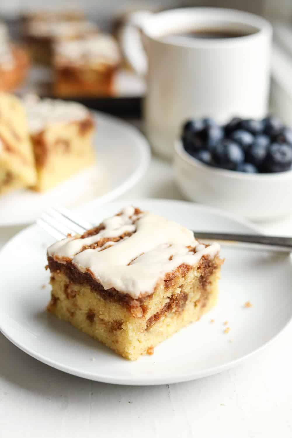 A piece of coffee cake on a white plate with a fork set by it. There's a coffee cup, blueberries, and more pieces of cake set behind the plate.