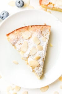 A piece of almond cake on a white plate with two blueberries next to it.