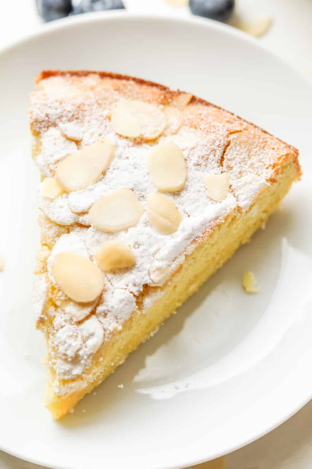 A slice of almond cake on a white plate with blueberries behind the plate.