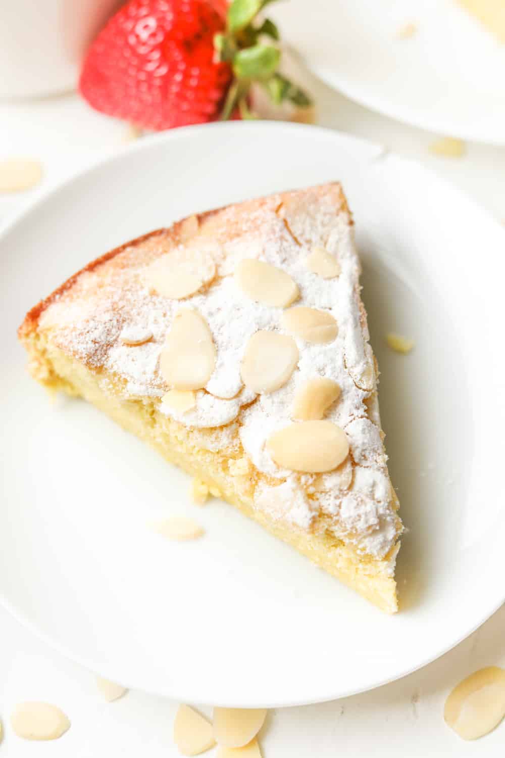 A piece of almond cake on a white plate with a strawberry next to the plate.