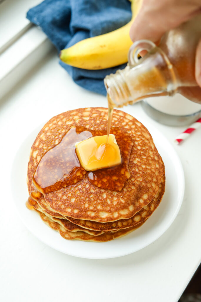 A stack of banana pancakes topped with butter and maple syrup being poured on them.