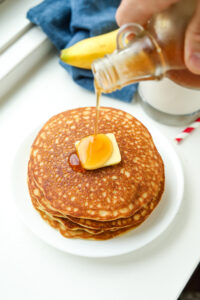 A stack of pancakes topped with butter and maple syrup is being poured on it.