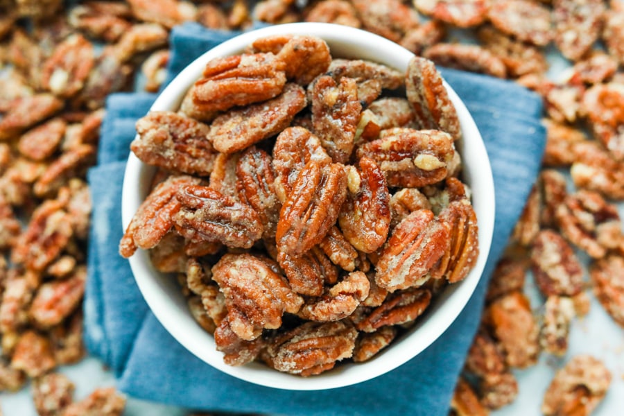 A white bowl filled with candied pecans. The bowl is set on a blue napkin and there are pecans all around the napkin.