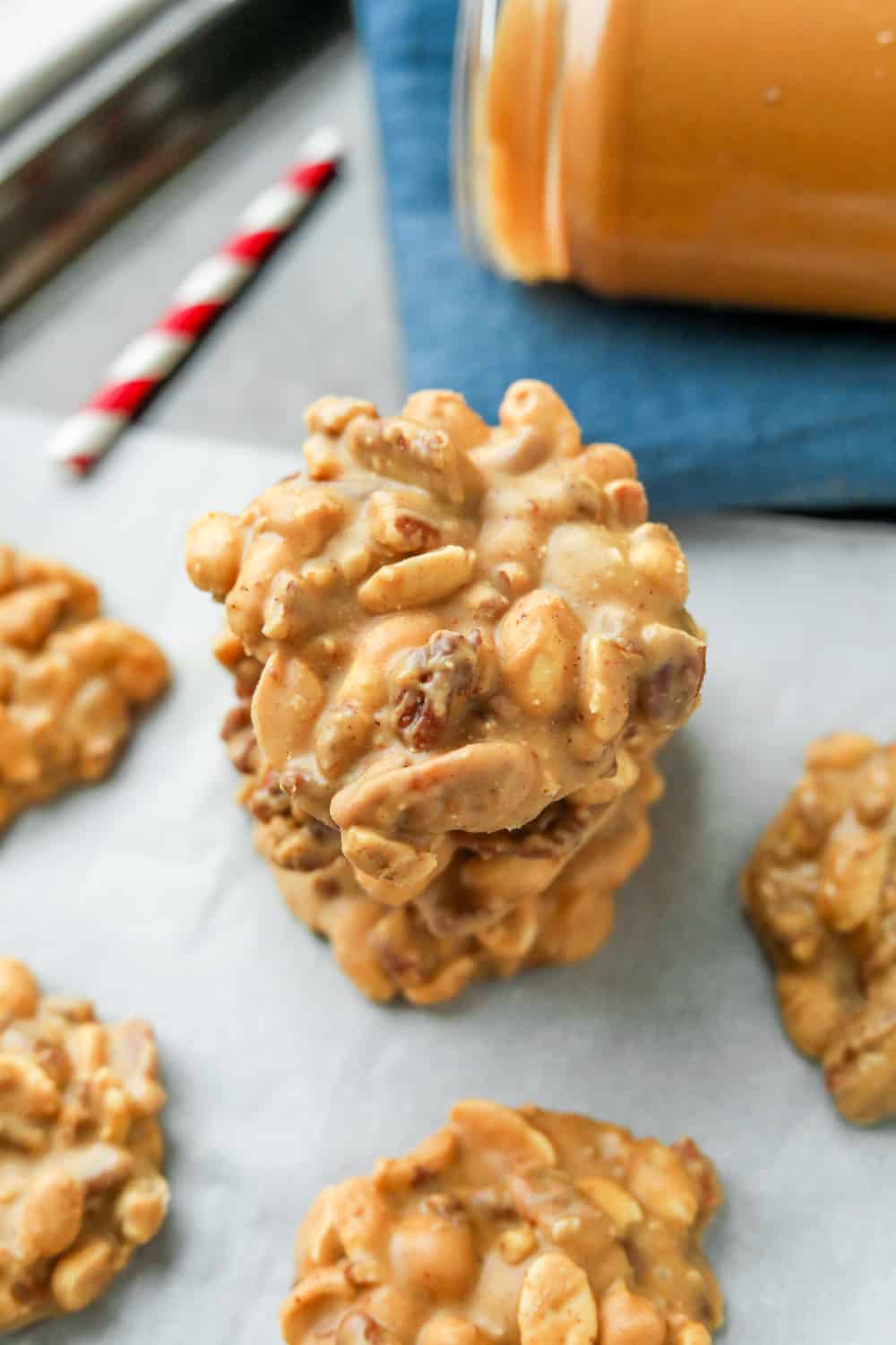 A stack of three peanut butter nut clusters on a sheet of white parchment paper with a blue towel and a red and white paper straw next to them.