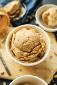 A white bowl with a scoop of peanut butter ice cream in it. The bowl is set on a cutting board, and behind it is a jar of peanut butter, a blue napkin, and another white bowl filled with ice cream.