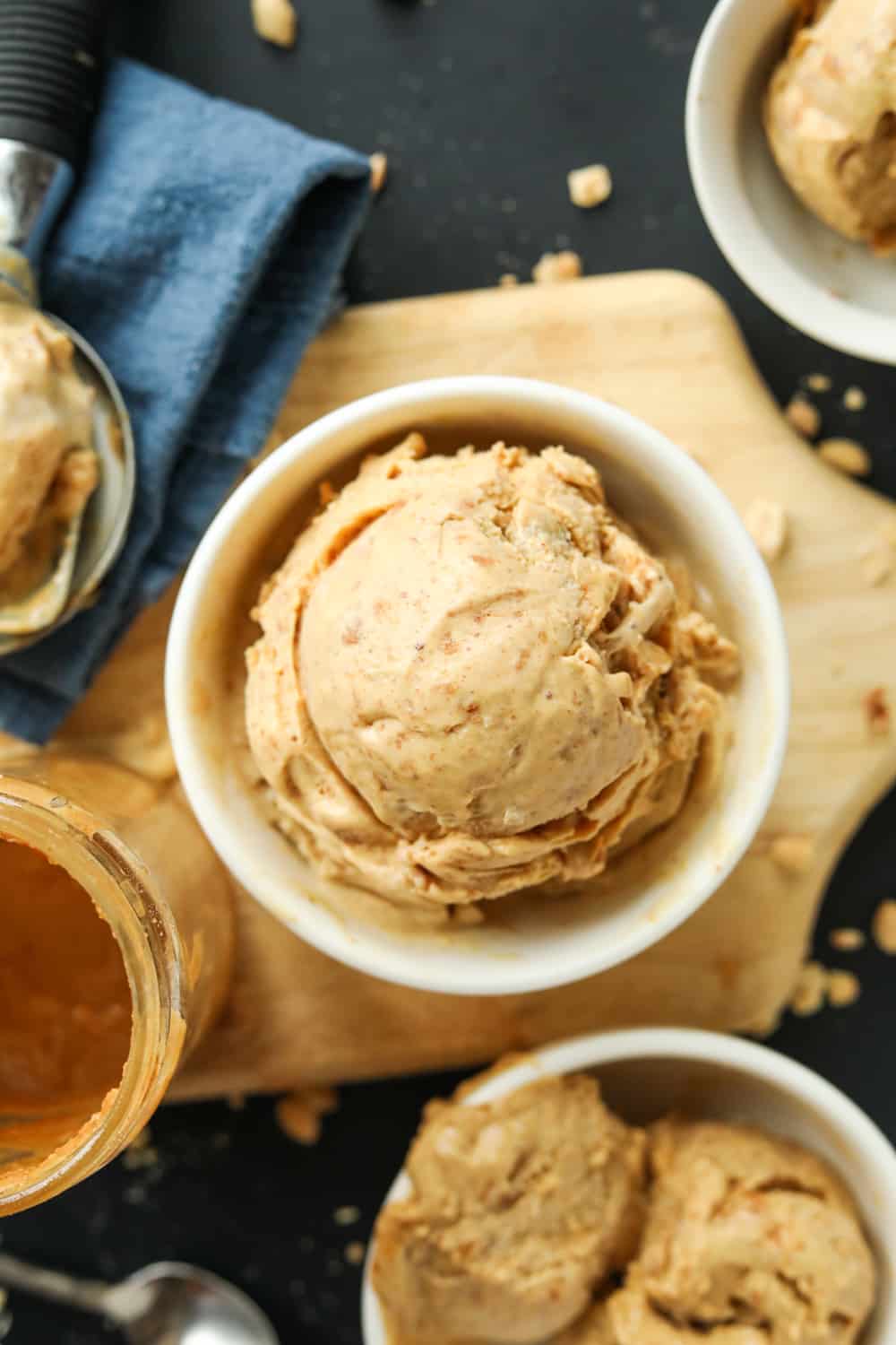 Three white bowls filled with peanut butter ice cream. One of the bowls is set on a wooden cutting board next to it is a blue napkin and a jar of peanut butter.