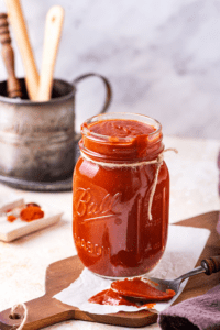 A glass jar filled with barbecue sauce. The jar is on a paper towel on a wooden cutting board and there is a spoon with barbecue sauce it on the paper towel.