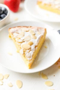 A slice of almond cake on a white plate. The plate is sat next to a bowl of blueberries, and there are strawberries next to the bowl.