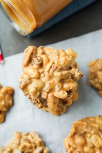 Nut clusters stacked on top of one another on a white sheet of parchment paper there is a jar of peanut butter next to them.