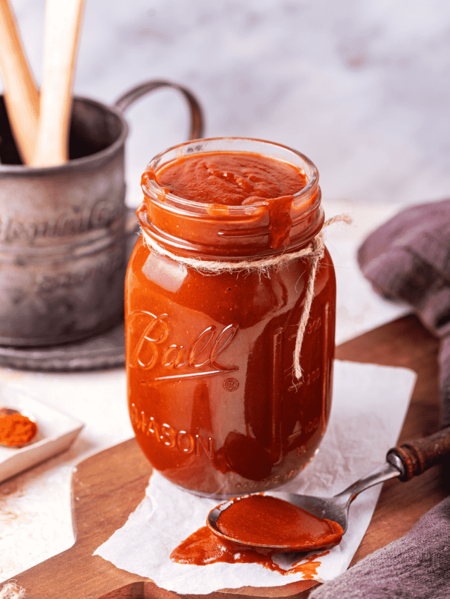 A glass jar filled with barbecue sauce on a paper towel on a wooden cutting board. There is a spoon with barbecue sauce on it on the paper towel.