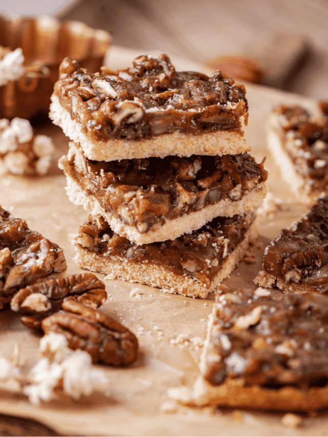 Three pecan pie bars stacked on top of one another on a piece of parchment paper. There are parts of three pecan pie bars to the right of the stack.