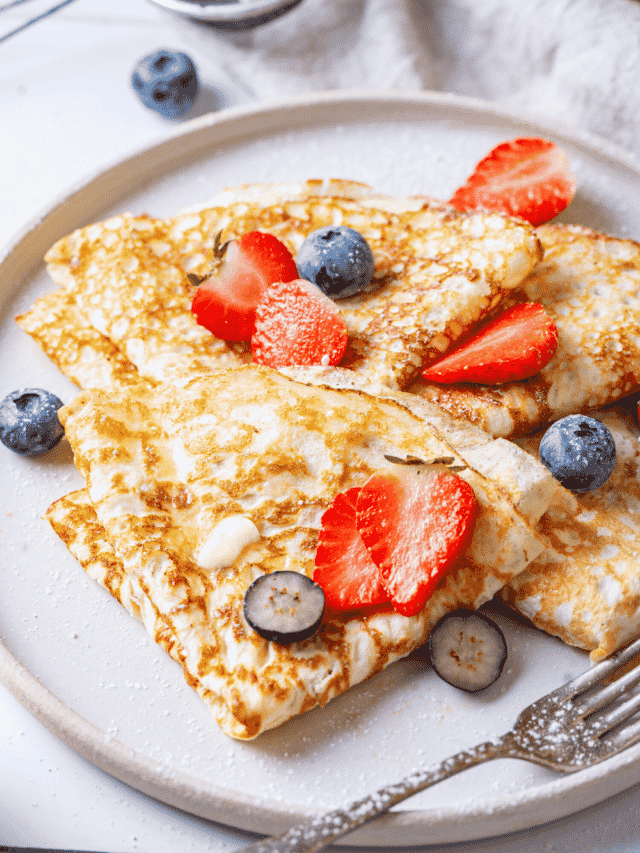 A white plate with a few crepes on it with some strawberry and blueberry slices on top.