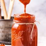 A glass jar filled to the top with barbecue sauce. A head of a spoon is dripping barbecue sauce from it into the top of the jar.