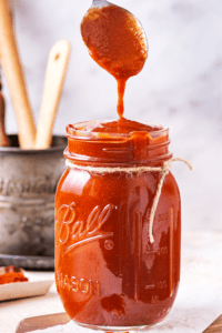 A glass jar filled to the top with barbecue sauce. A head of a spoon is dripping barbecue sauce from it into the top of the jar.