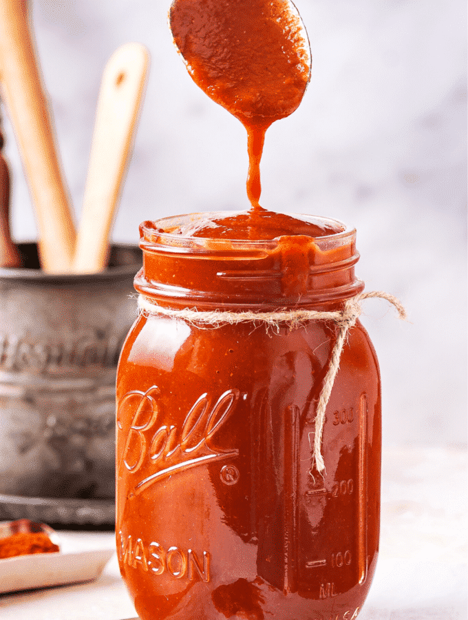 A glass jar filled to the top with barbecue sauce. A head of a spoon is dripping barbecue sauce from it into the top of the jar.