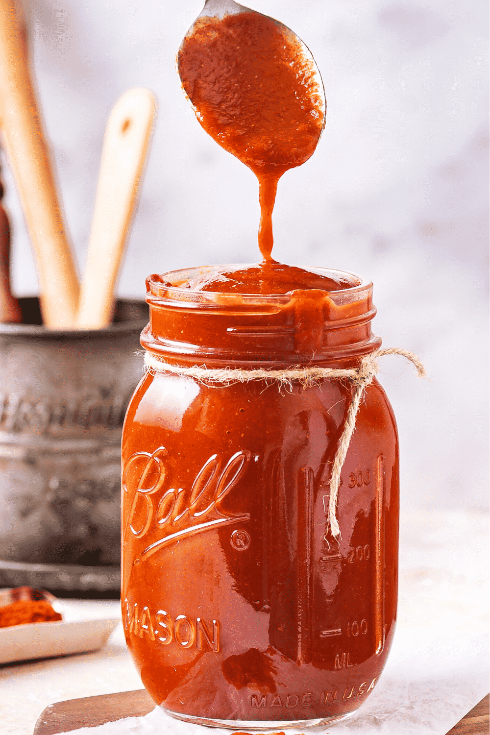 A glass jar filled to the top with BBQ sauce. A head of a spoon is dripping BBQ sauce from it into the top of the jar.