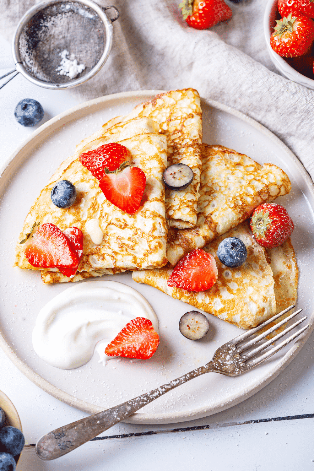 A white plate with for crêpes overlapping one another on it. There are slices of strawberries and blueberries on the crepes and a fork on the plate.