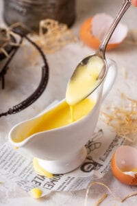 A gravy boat filled with hollandaise sauce. A spoon is being held over the gravy boat and hollandaise is dripping from the spoon into the boat.